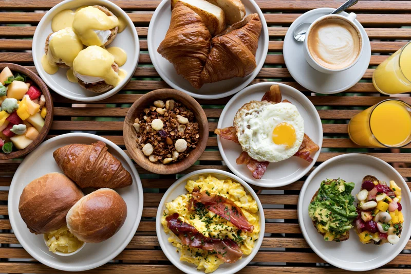 Overhead view of a full brunch spread with croissants, eggs, avocado toast, granola, fruit, coffee and orange juice on a slatted wooden table