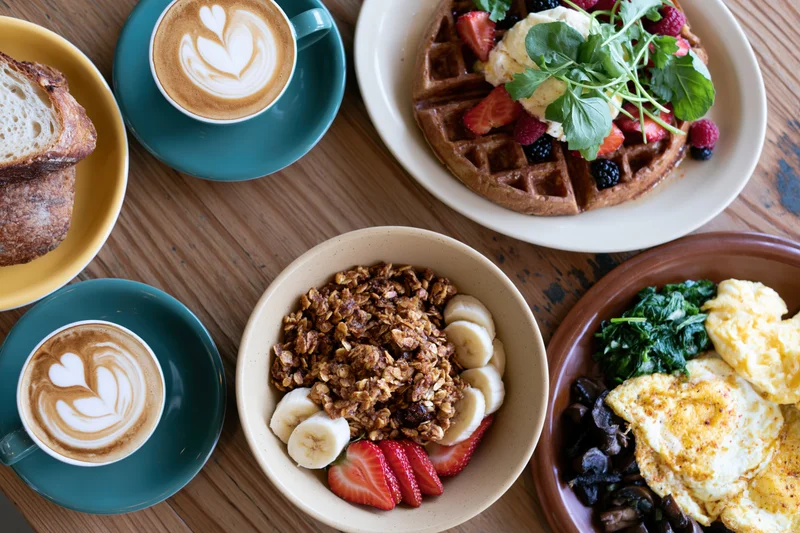 Overhead flat lay of a cafe brunch spread with coffee, waffles, granola, eggs and croissant on a wooden table