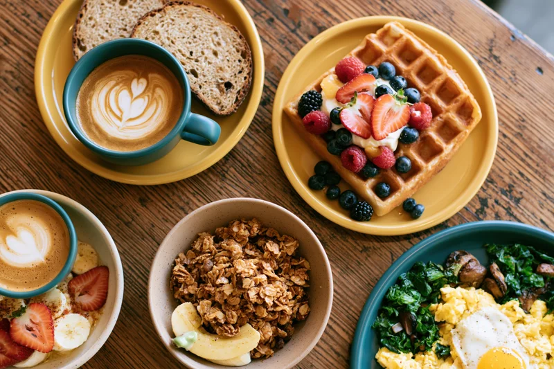 Overhead flat lay of a cafe brunch spread with waffles, coffee, granola, and eggs on a wooden table