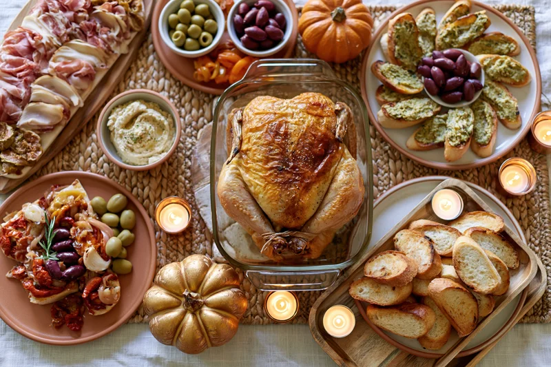 Roasted turkey in a glass dish surrounded by bread, olives, cheese, charcuterie, and pumpkins on a woven placemat with candles