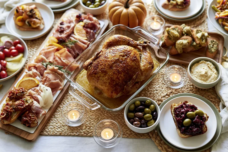 Roasted turkey on a glass platter surrounded by charcuterie, bread, olives, and candles on a thanksgiving dinner table