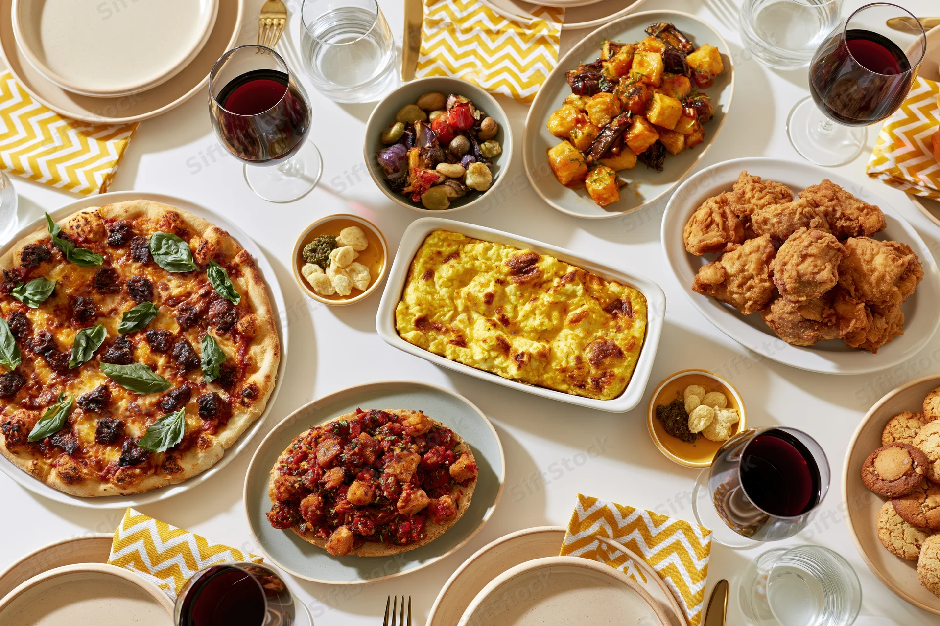 Overhead view of a fully set dining table with pizza, fried chicken, pasta, vegetables, wine, and desserts arranged for a dinner party