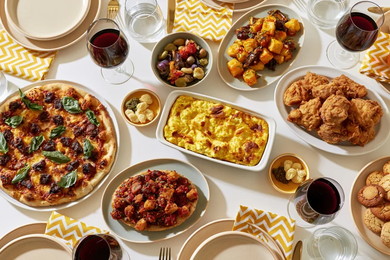 Overhead view of a fully set dining table with pizza, fried chicken, pasta, vegetables, wine, and desserts arranged for a dinner party