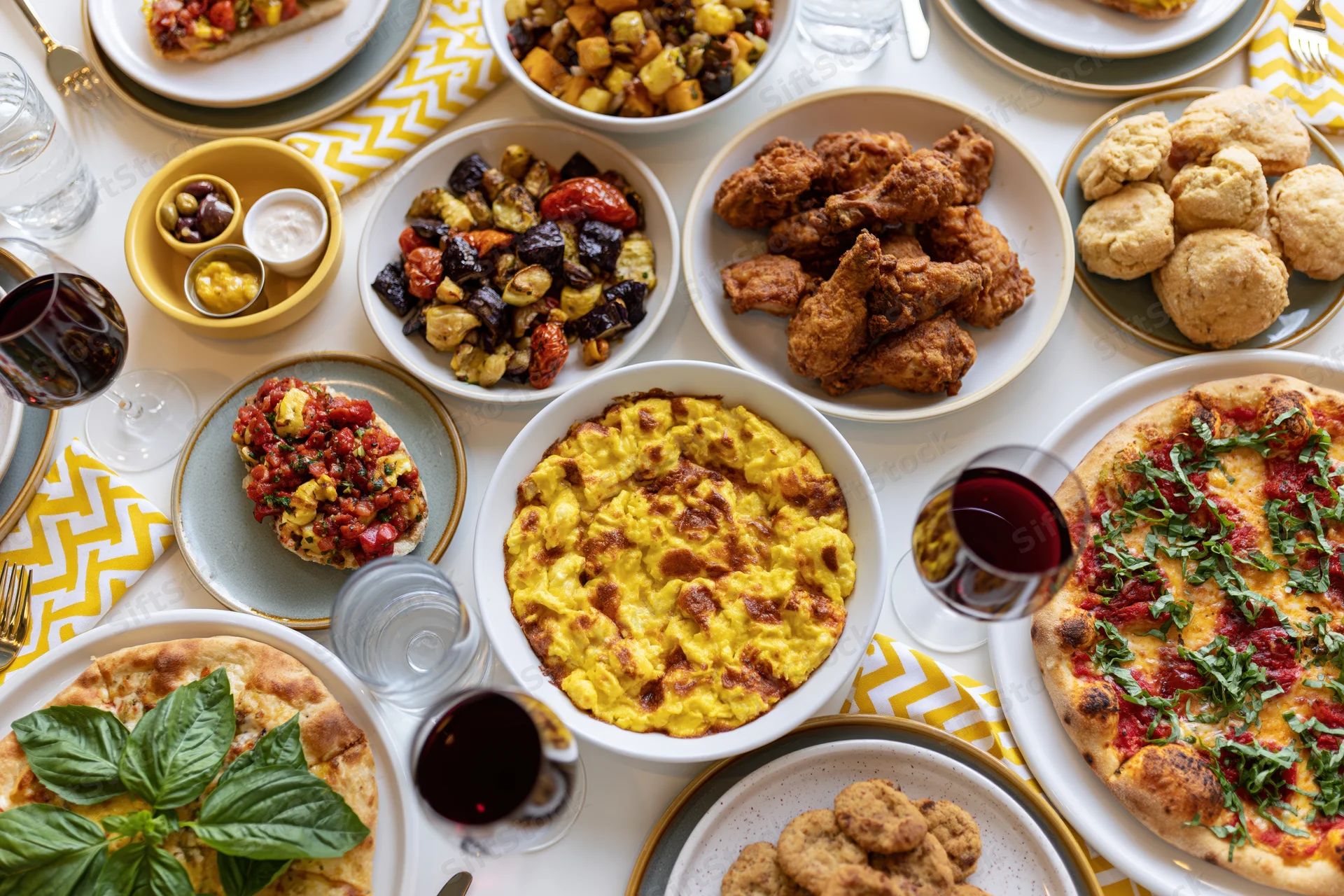 Overhead view of a fully set dining table with pizza, fried chicken, pasta, vegetables, biscuits, and wine glasses arranged for a shared meal