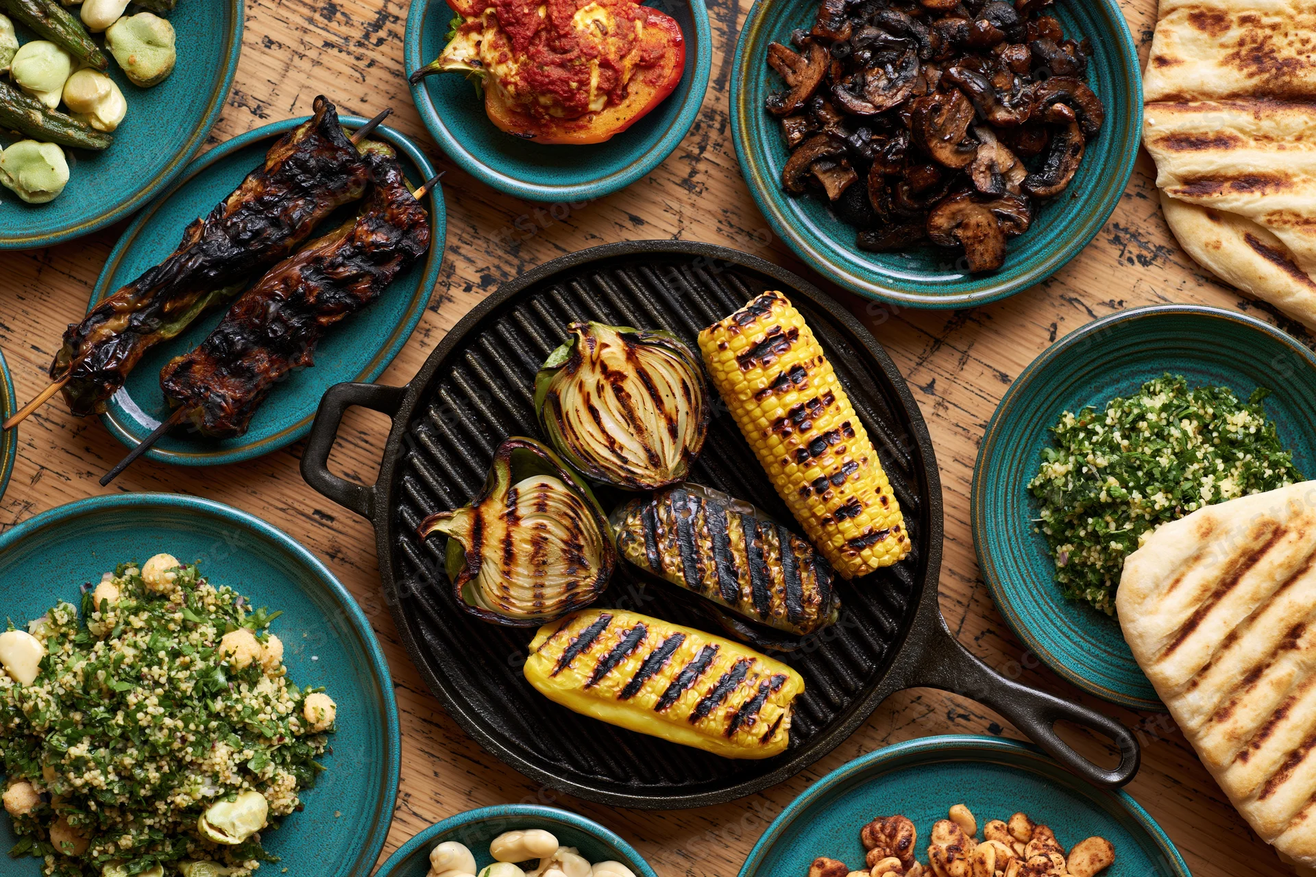 Grilled vegetables and side dishes arranged in a flat lay on a wooden table with cast iron skillet and teal plates