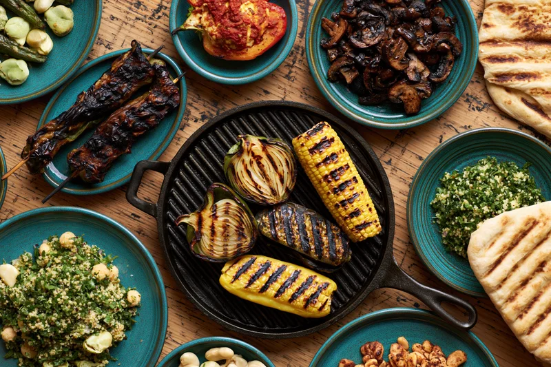 Grilled vegetables and side dishes arranged in a flat lay on a wooden table with cast iron skillet and teal plates