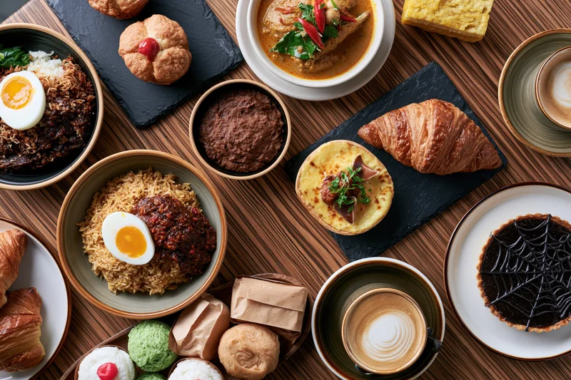 Overhead flat lay of a southeast asian cafe breakfast spread on a wooden table with rice dishes, pastries, coffee and themed desserts