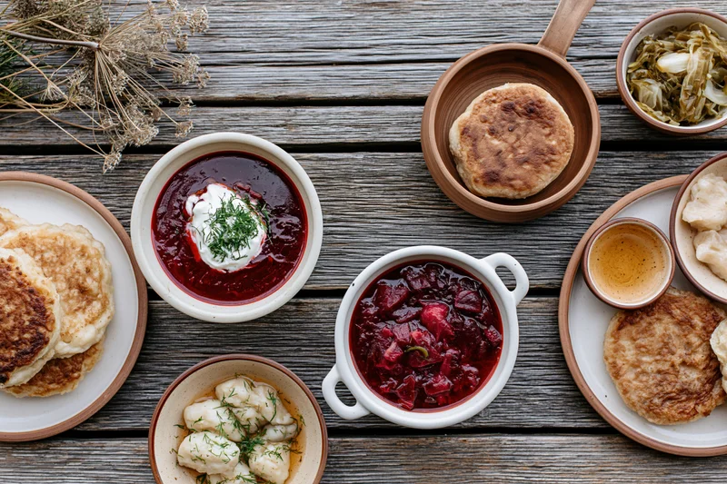Overhead flat lay of a rustic eastern european meal spread on a wooden table featuring borscht, pierogi, blini, and sauerkraut
