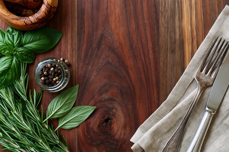 Overhead flat lay of a rustic dark walnut wooden table with fresh herbs, peppercorns, and silverware on a linen napkin