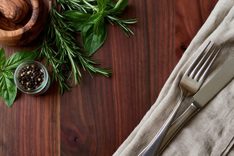 Overhead flat lay of rustic dark walnut wooden table with cutlery, herbs, pepper, and mortar on linen napkin