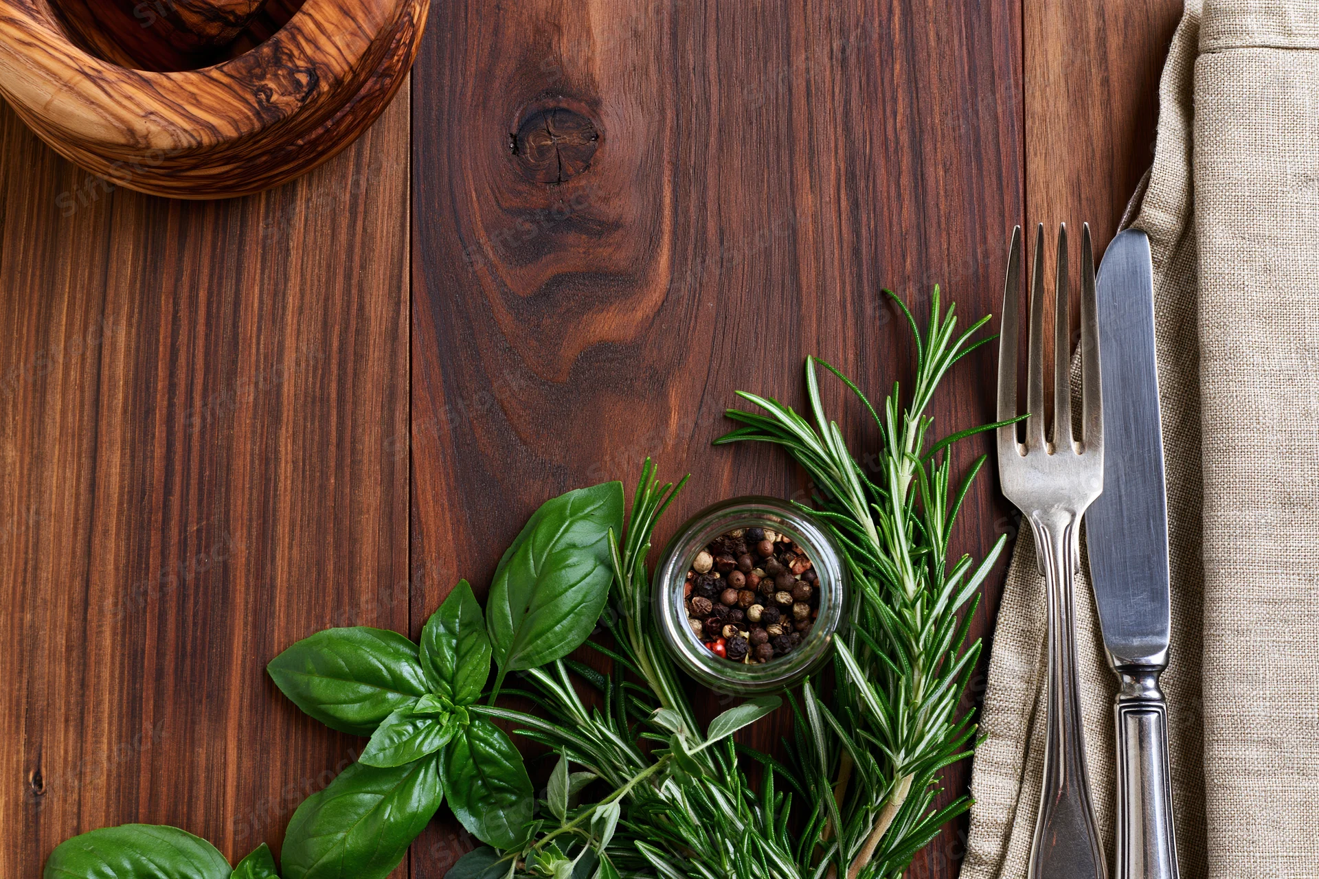 Overhead flat lay of fresh herbs, spices, and cutlery arranged on a dark walnut wooden table surface