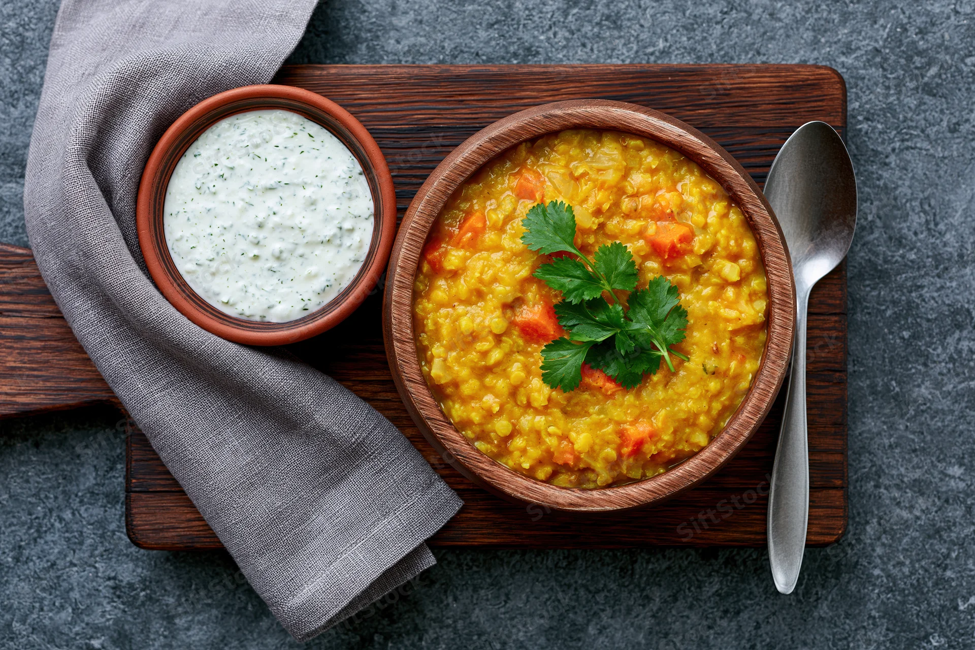Yellow lentil dal with cilantro garnish and yogurt raita side in wooden bowls on dark stone surface
