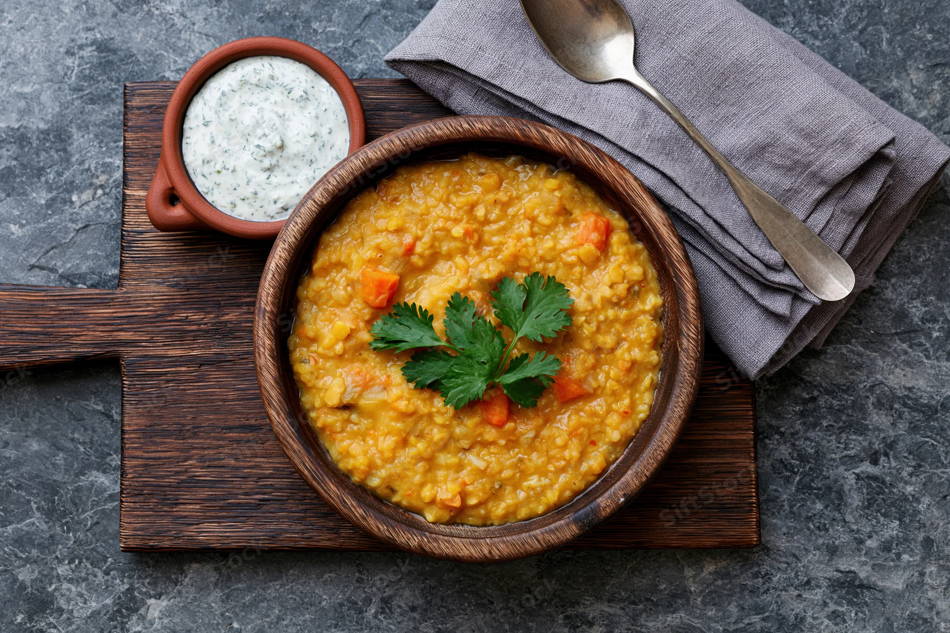Wooden bowl of yellow lentil dal with cilantro garnish and yogurt raita on a cutting board