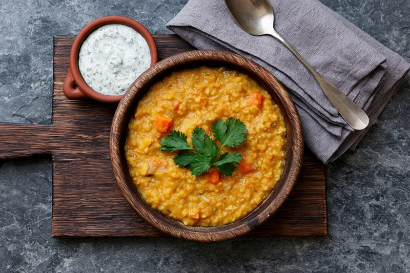 Wooden bowl of yellow lentil dal with cilantro garnish and yogurt raita on a cutting board