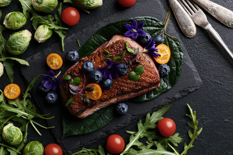 Seared duck breast fillet garnished with blueberries, kumquats, and edible flowers on a slate platter surrounded by arugula and brussels sprouts