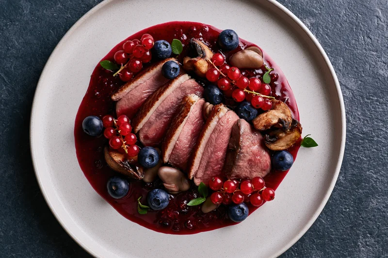 Sliced medium-rare duck breast with berries and mushrooms on a white plate over dark slate surface