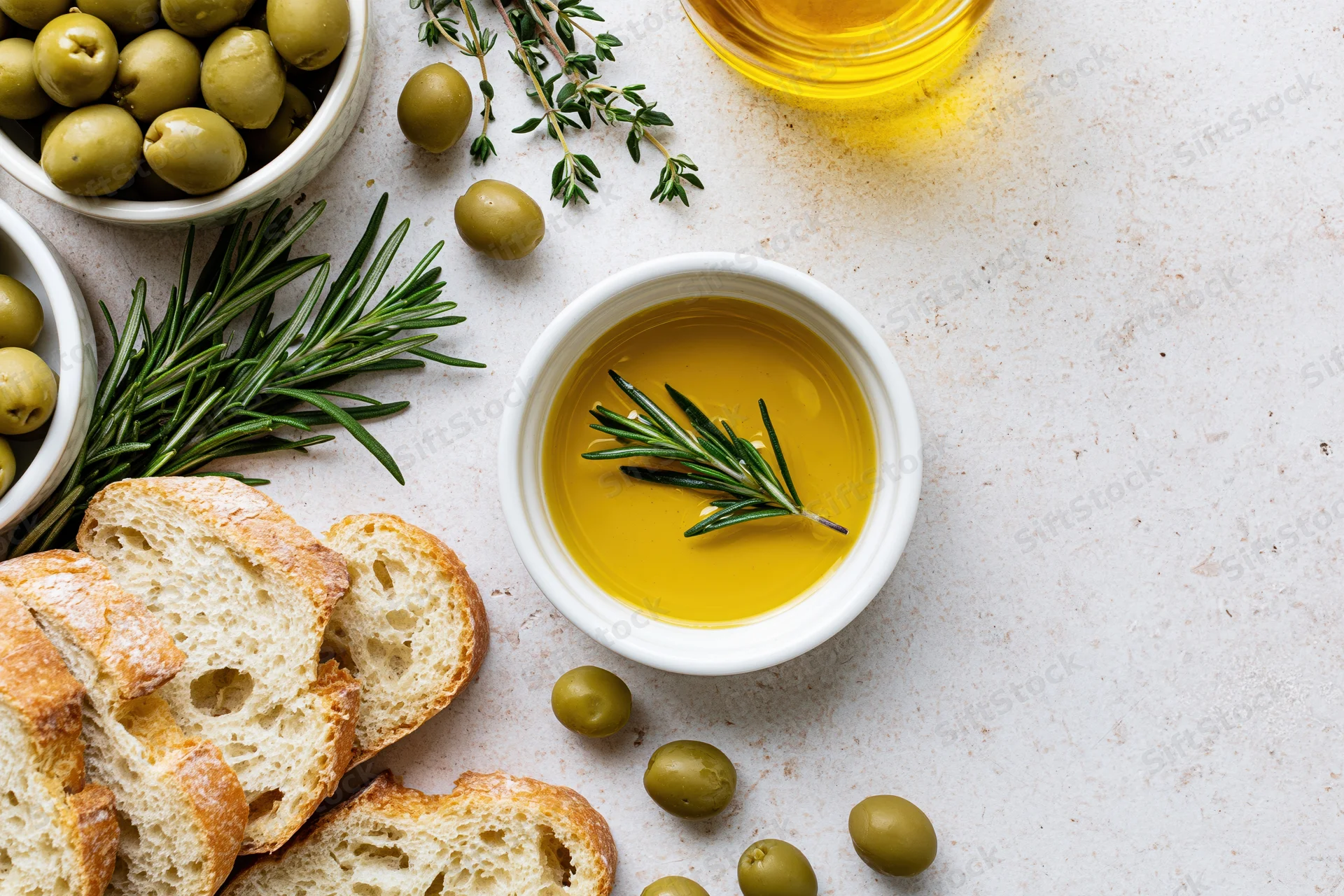 Overhead flat lay of olive oil in a white ceramic bowl with bread, olives, and herbs on a light surface