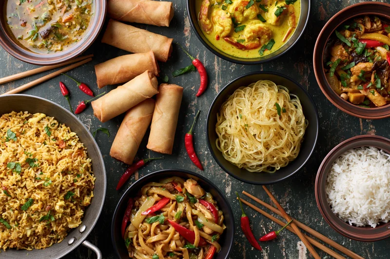 Overhead flat lay of various asian dishes including fried rice, spring rolls, curry, noodles, and stir fry arranged on a dark wooden table with chopsticks and red chilies scattered around