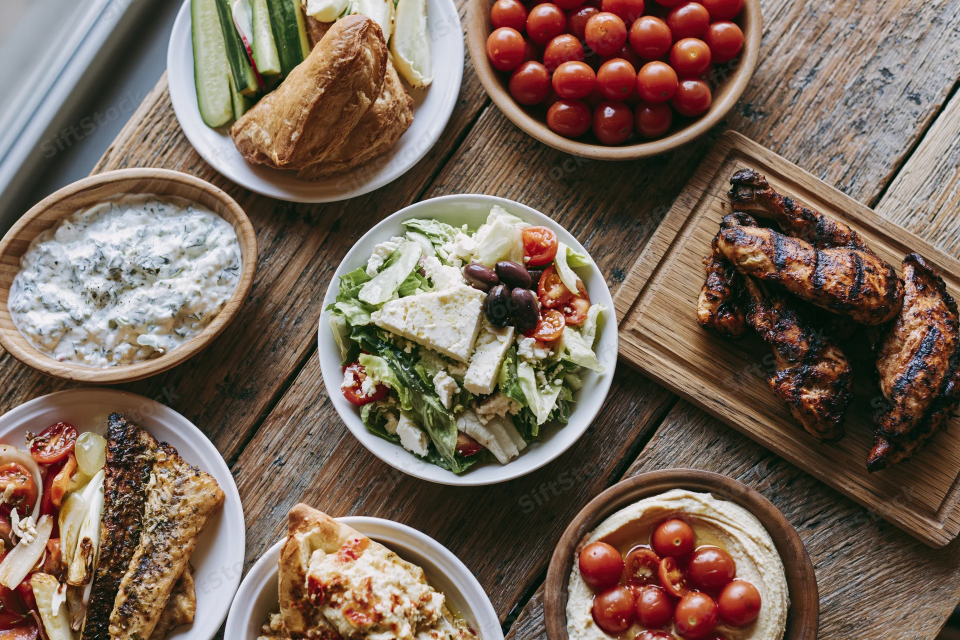 Overhead view of greek mezze spread with grilled chicken, fish, salad, hummus, tzatziki, and vegetables on a wooden table