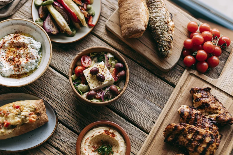 Overhead flat lay of greek mezze dishes arranged on a wooden table with bread, hummus, tzatziki, grilled chicken and vegetables