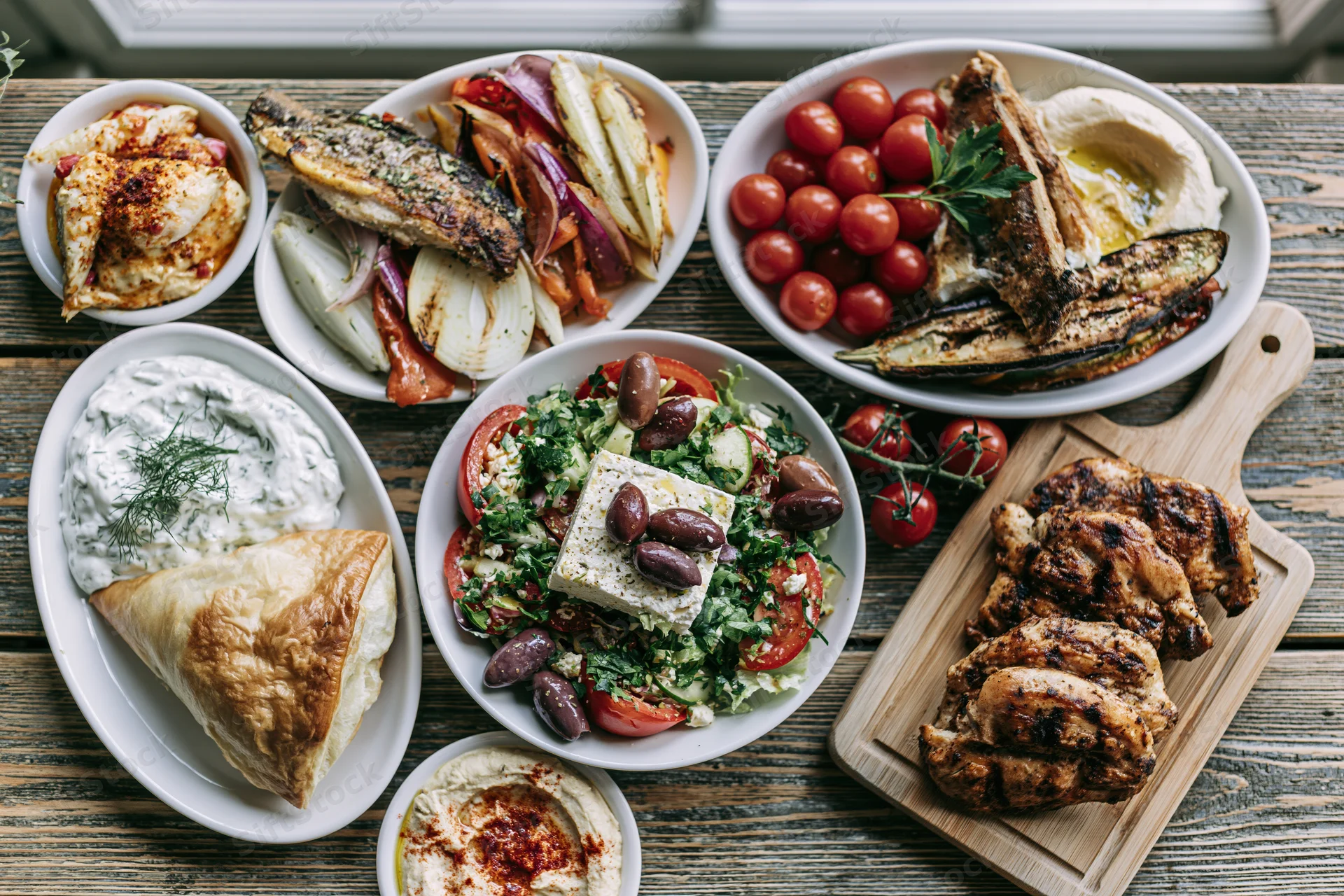 Overhead view of greek mezze dishes arranged on a wooden table with grilled chicken, fish, salad, hummus and tzatziki