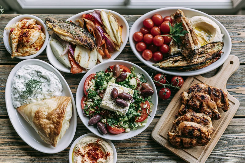 Overhead view of greek mezze dishes arranged on a wooden table with grilled chicken, fish, salad, hummus and tzatziki