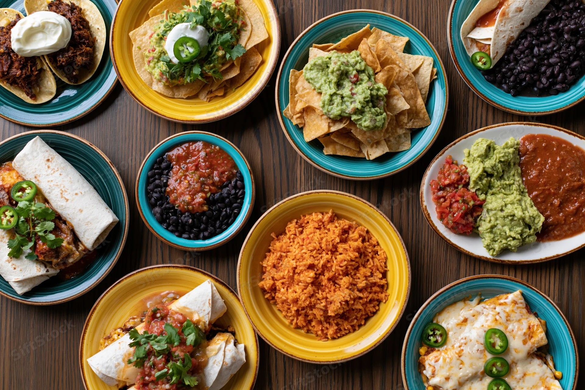 Overhead flat lay of mexican dishes spread on wooden table including burritos, tacos, enchiladas, rice, beans, guacamole, salsa, and tortilla chips on colorful plates
