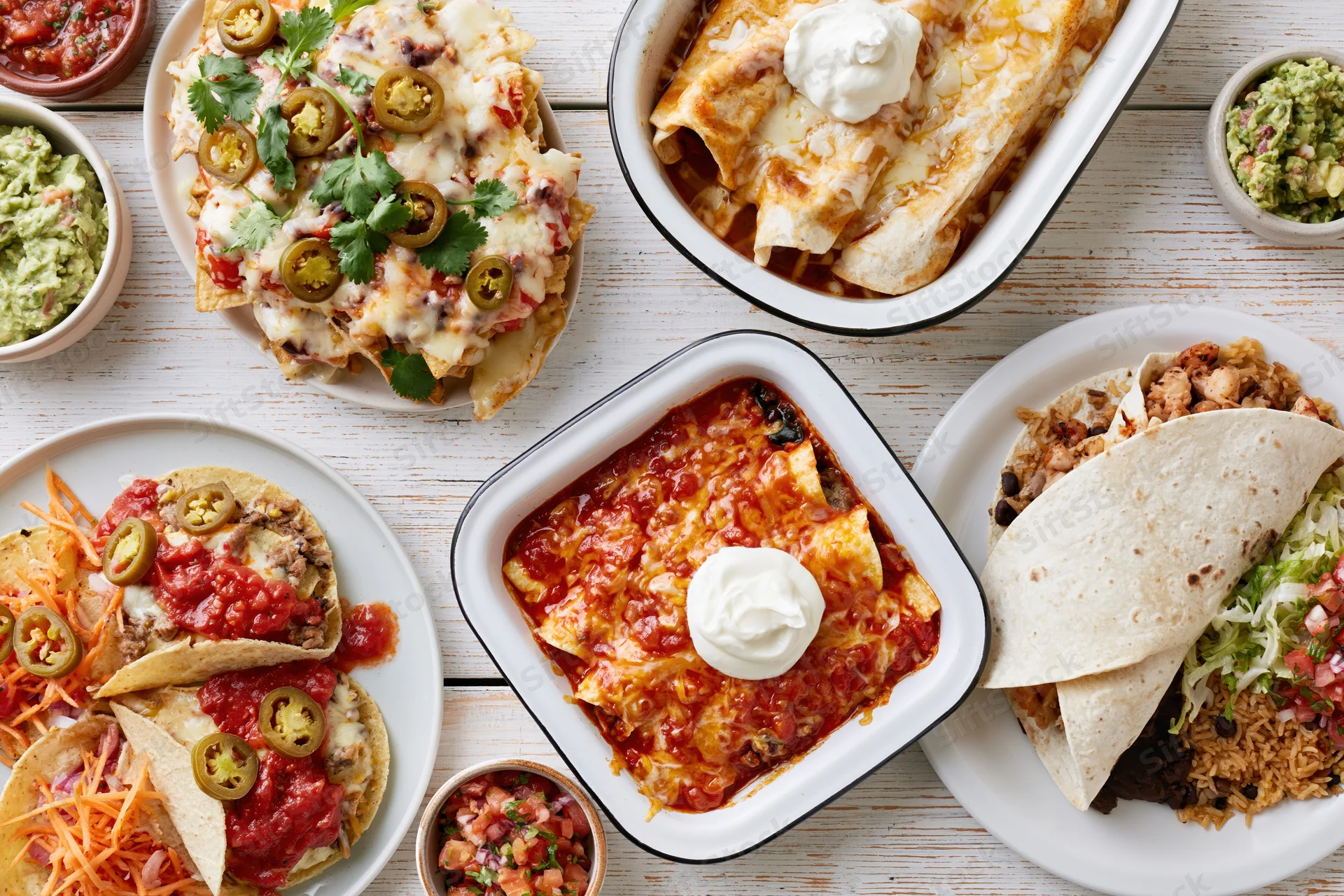 Overhead flat lay of mexican dishes including tacos, enchiladas, burritos, nachos, and sides arranged on a white wooden table