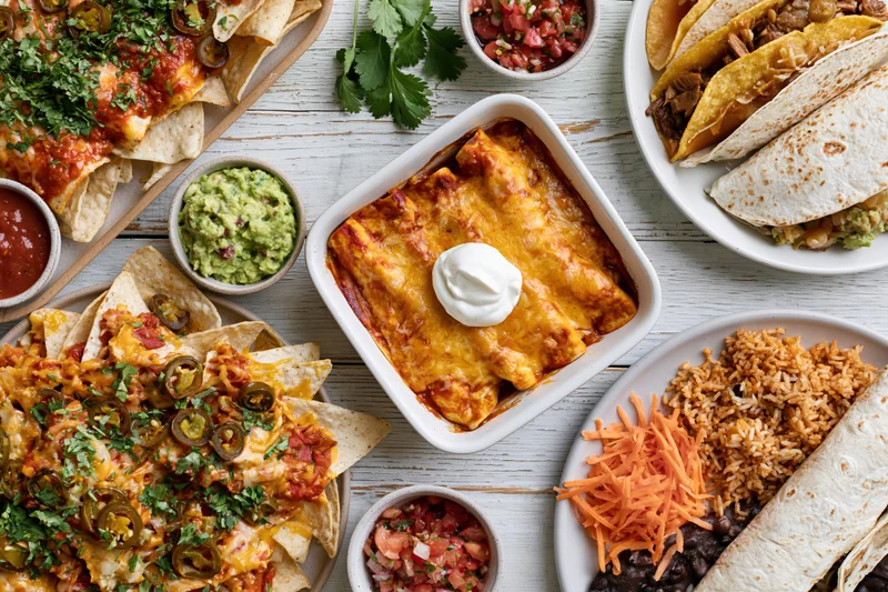 Overhead flat lay of mexican dishes including enchiladas, nachos, tacos, and rice on a white wooden table