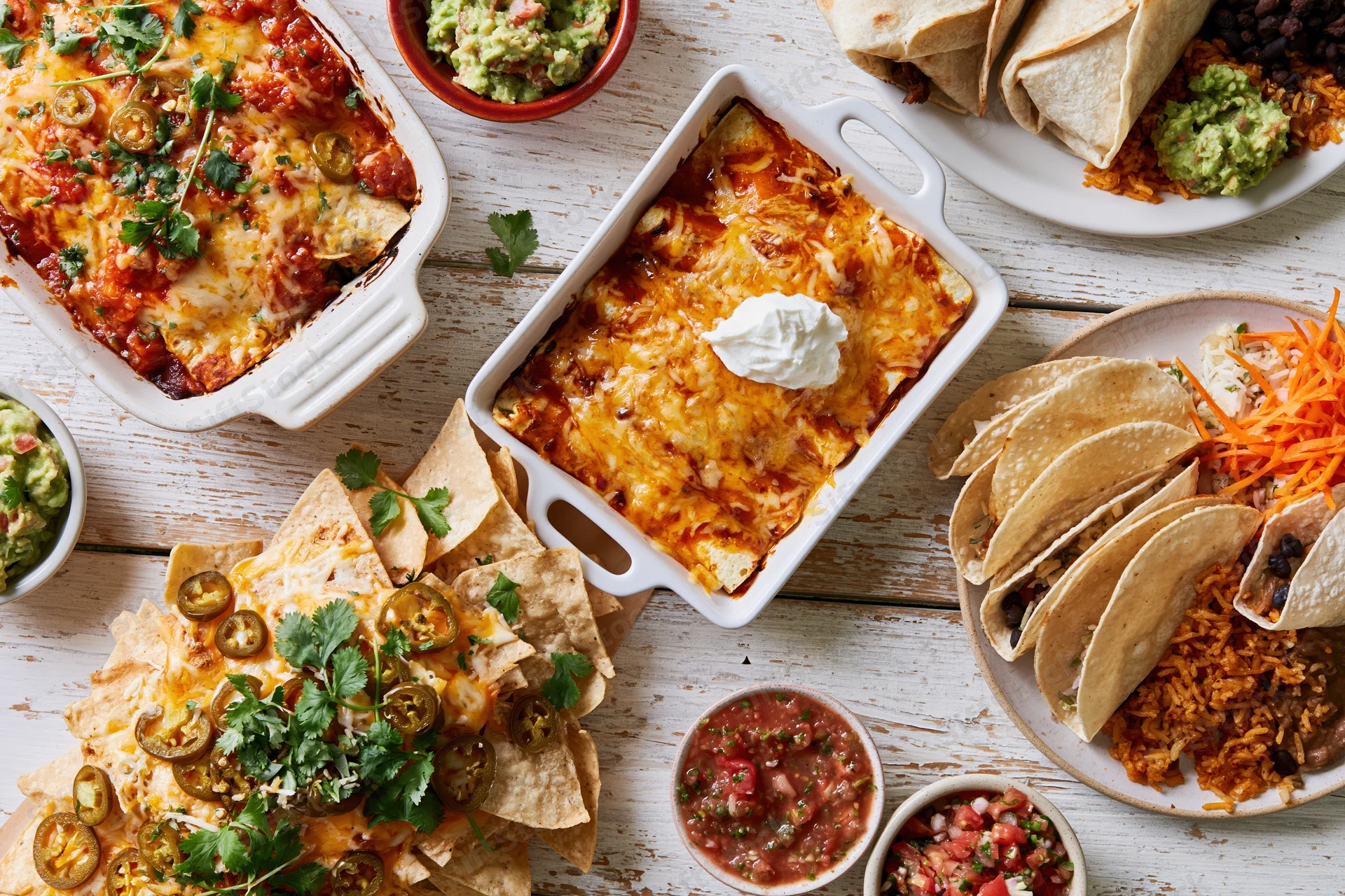 Overhead flat lay of mexican dishes including enchiladas, nachos, tacos, and guacamole on a white wooden table