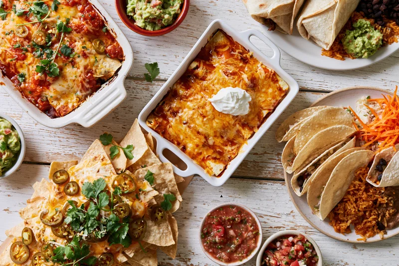 Overhead flat lay of mexican dishes including enchiladas, nachos, tacos, and guacamole on a white wooden table