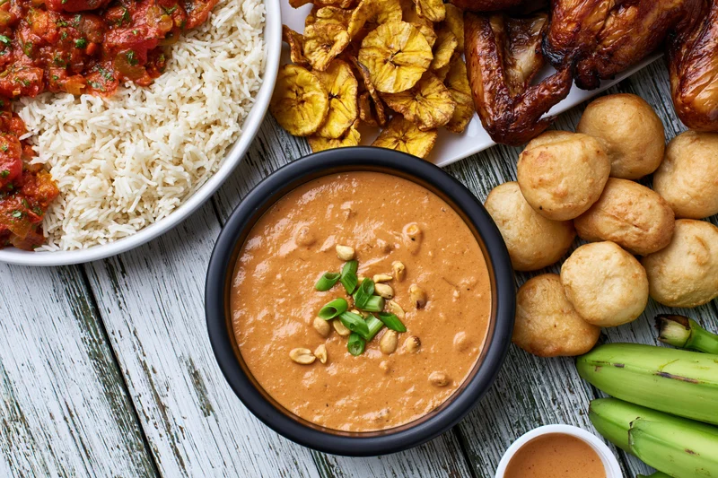Overhead flat lay of a West African meal spread on a weathered wooden table featuring rice, peanut stew, fried plantains, chicken, dumplings, and green bananas