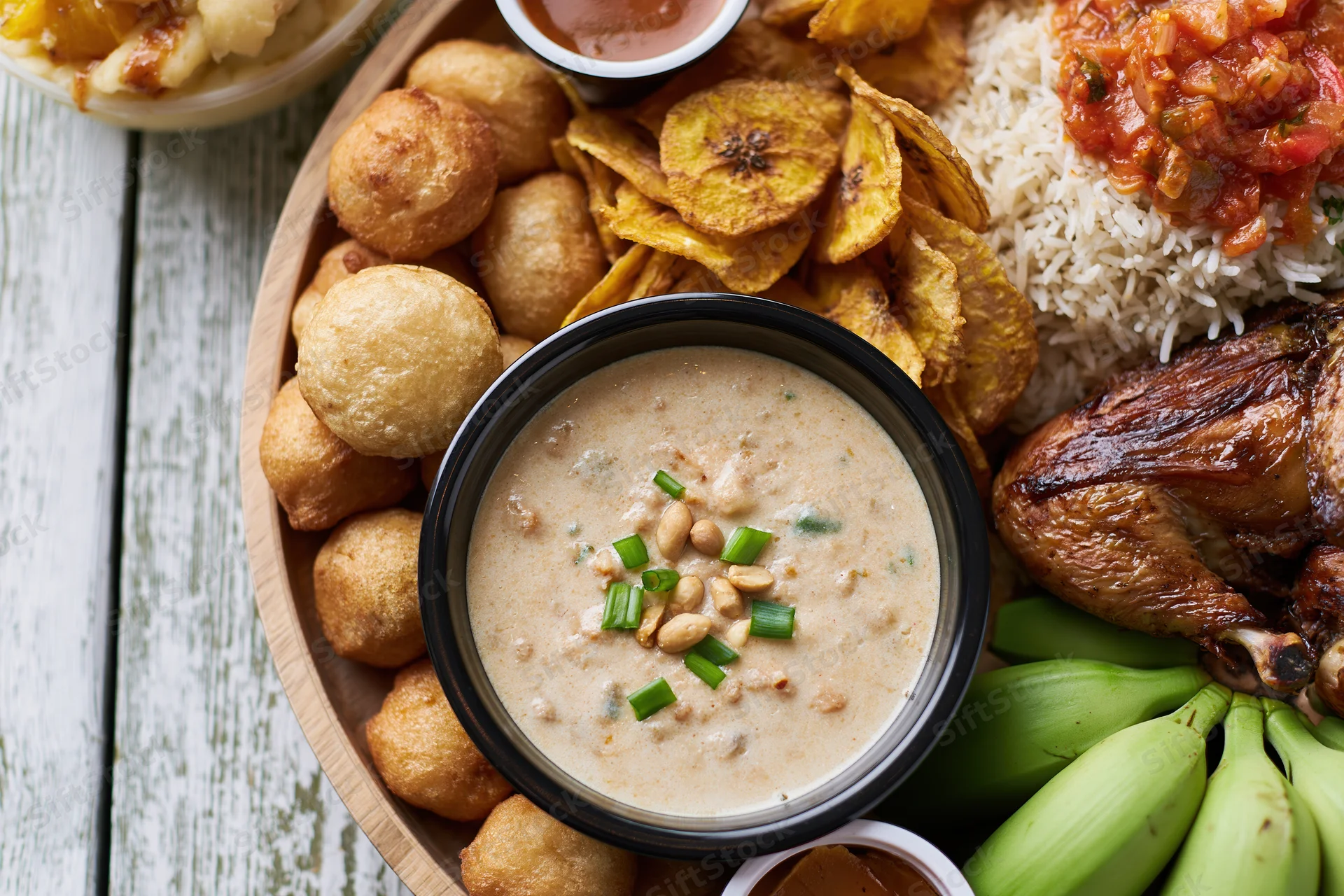 Overhead view of a West African meal spread with rice, fried chicken, plantain, fufu, and peanut soup on a wooden table