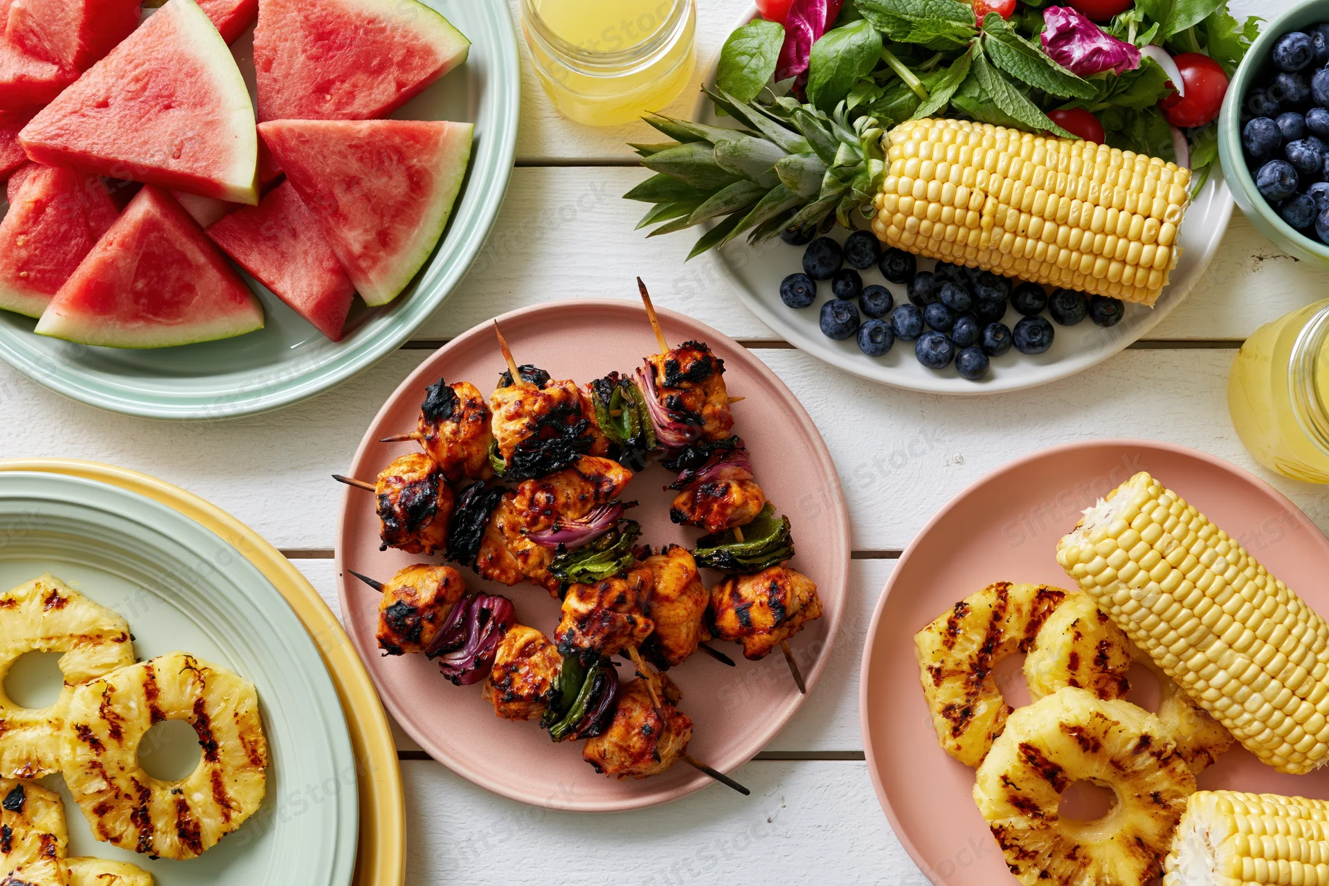 Overhead flat lay of a summer barbecue spread with grilled chicken skewers, corn, pineapple, watermelon, and salad on a white wooden table