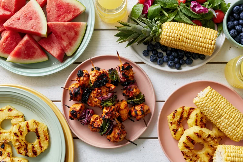 Overhead flat lay of a summer barbecue spread with grilled chicken skewers, corn, pineapple, watermelon, and salad on a white wooden table