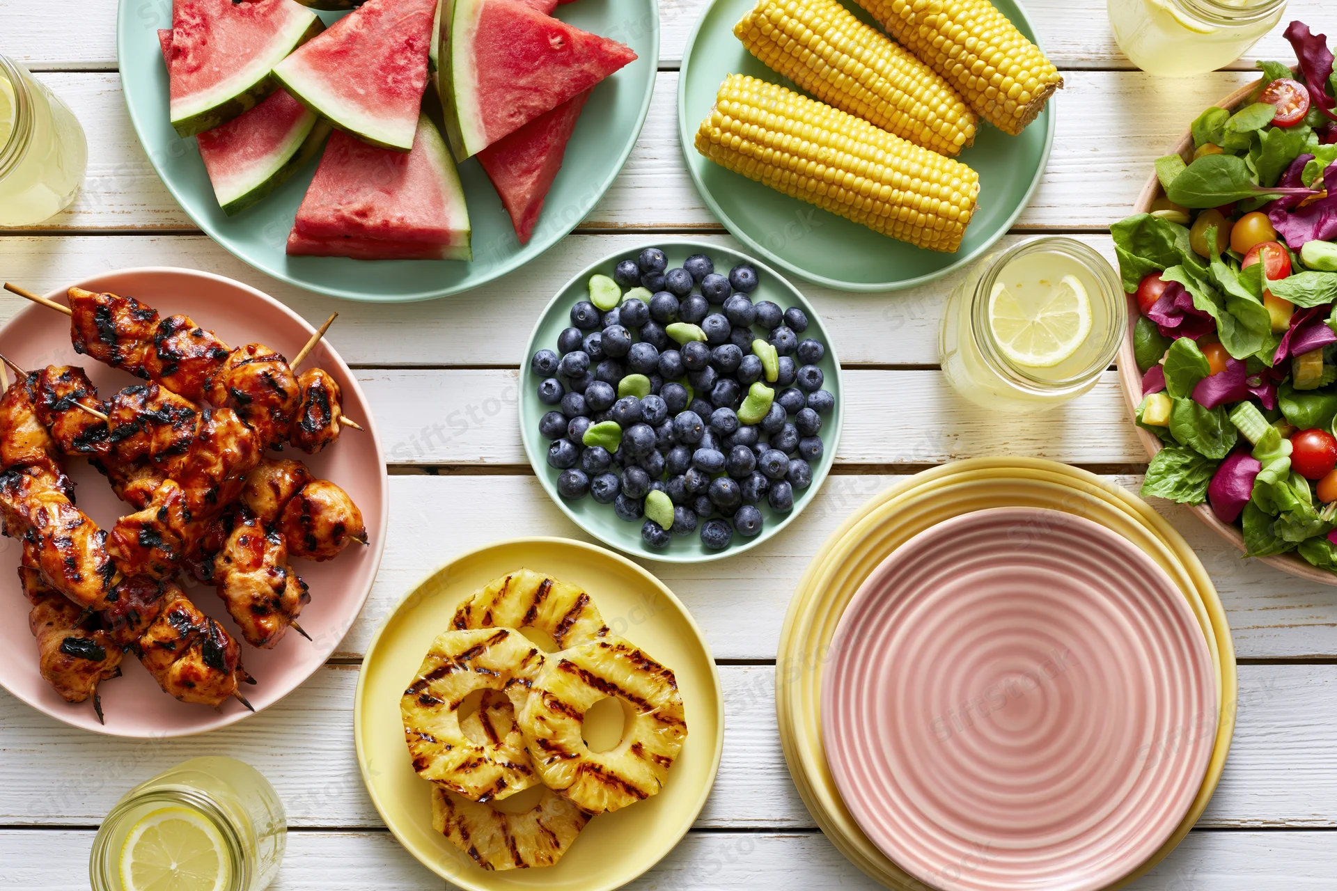 Overhead flat lay of a summer barbecue spread with grilled chicken, corn, watermelon, blueberries, pineapple, salad, and lemonade on a white wooden table