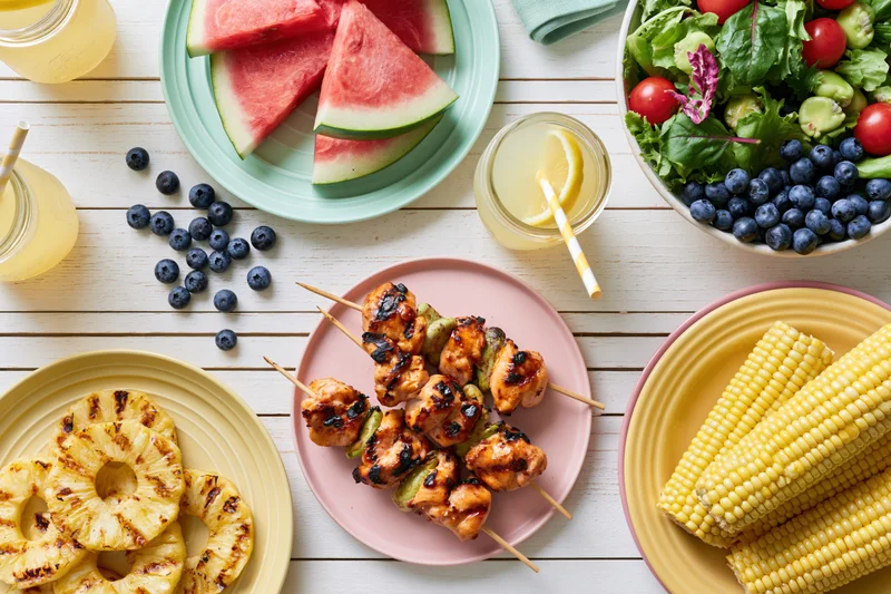 Overhead flat lay of a summer barbecue spread with grilled chicken skewers, corn, pineapple, watermelon, salad and drinks on a white wooden table