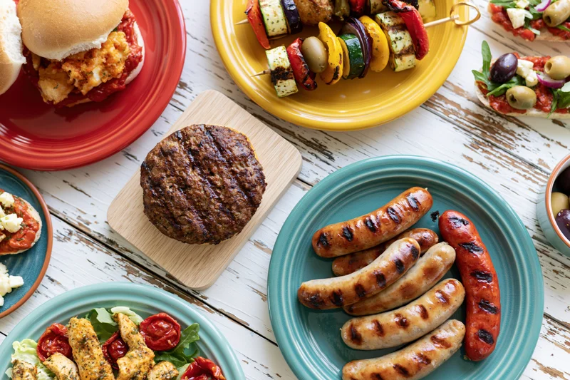 Grilled sausages, burger, and vegetable kebabs arranged on colorful plates on a white wooden table