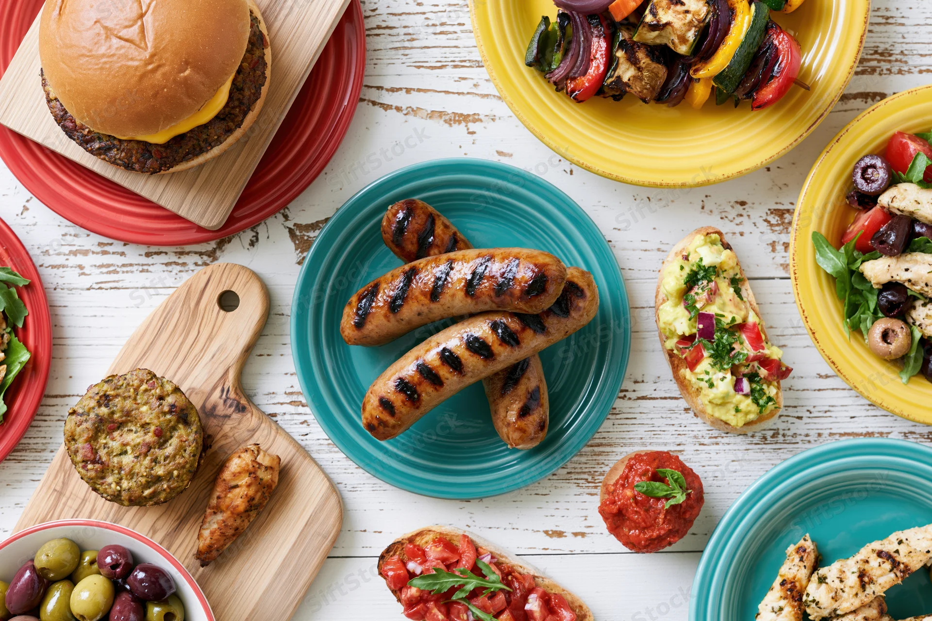 Overhead flat lay of grilled sausages, burgers, vegetables and salads on a white wooden table with colorful plates and cutting boards
