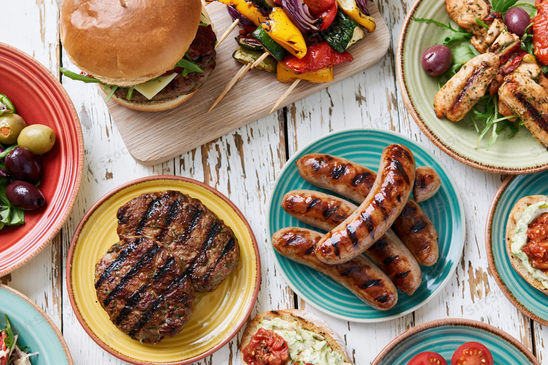 Overhead view of grilled sausages, burgers, kebabs and vegetables arranged on colorful plates on a white wooden table