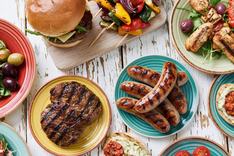 Overhead view of grilled sausages, burgers, kebabs and vegetables arranged on colorful plates on a white wooden table