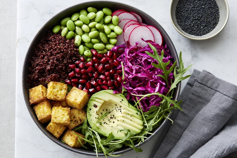 Overhead flat lay of a vegan buddha bowl with red quinoa, avocado, tofu, edamame, radish, purple cabbage, pomegranate, arugula, and black seeds on a marble surface