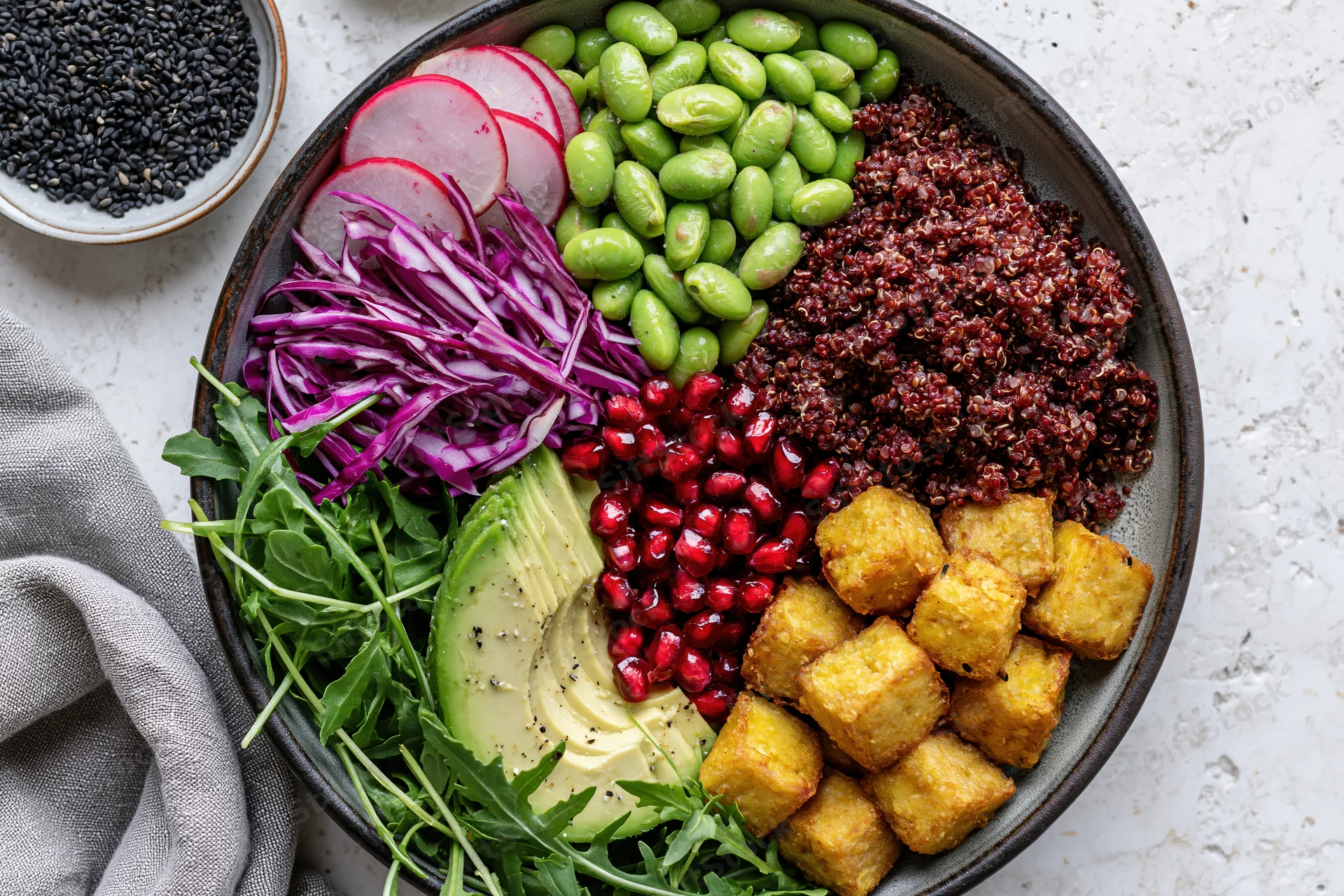 Overhead flat lay of a vegan buddha bowl with red quinoa, edamame, avocado, pomegranate, tofu, radish, cabbage, arugula, and black sesame on a textured surface