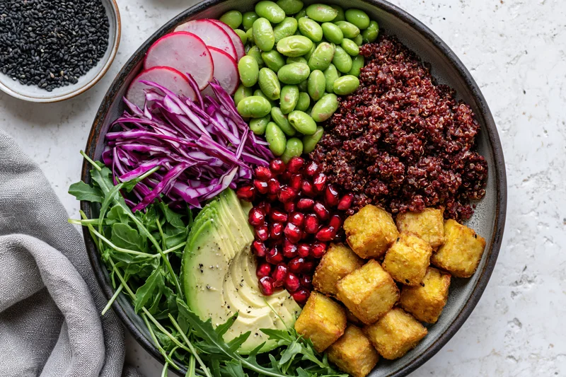 Overhead flat lay of a vegan buddha bowl with red quinoa, edamame, avocado, pomegranate, tofu, radish, cabbage, arugula, and black sesame on a textured surface