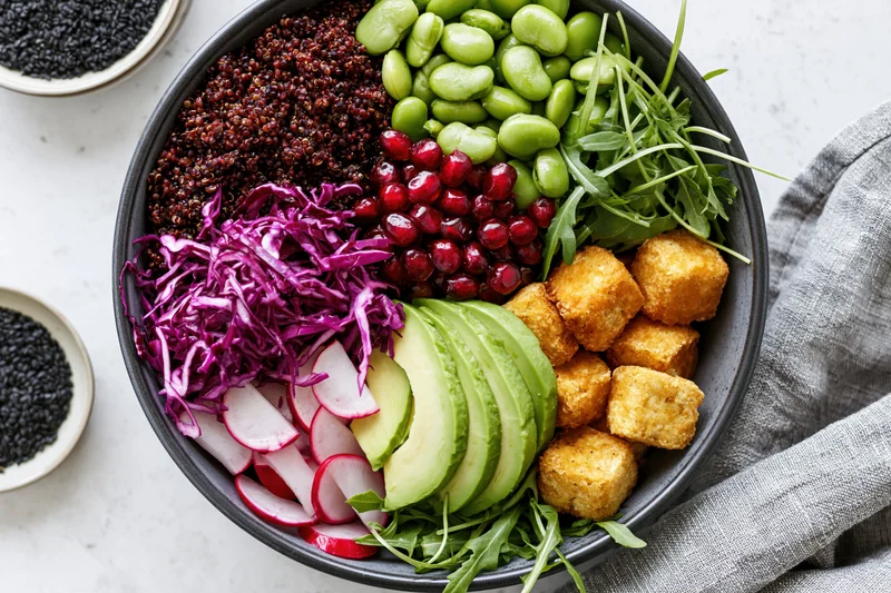 Overhead view of a vegan buddha bowl filled with red quinoa, avocado, tofu, edamame, pomegranate, radish, red cabbage, arugula, and black sesame on a white countertop with a gray linen napkin