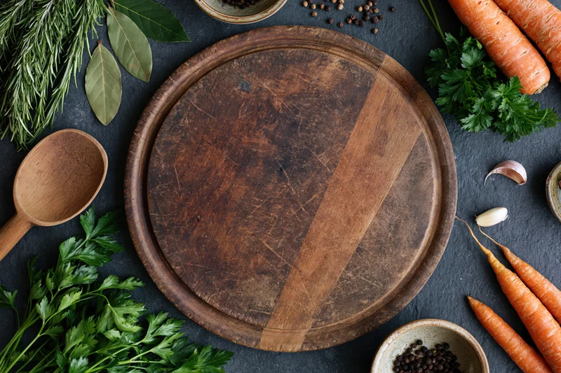 Round wooden cutting board surrounded by fresh herbs and vegetables on dark surface