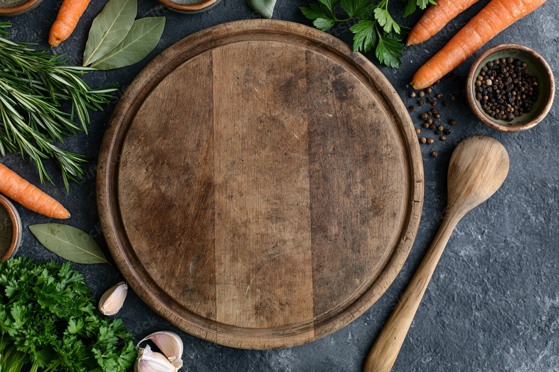 Round wooden cutting board surrounded by fresh vegetables and herbs on dark surface