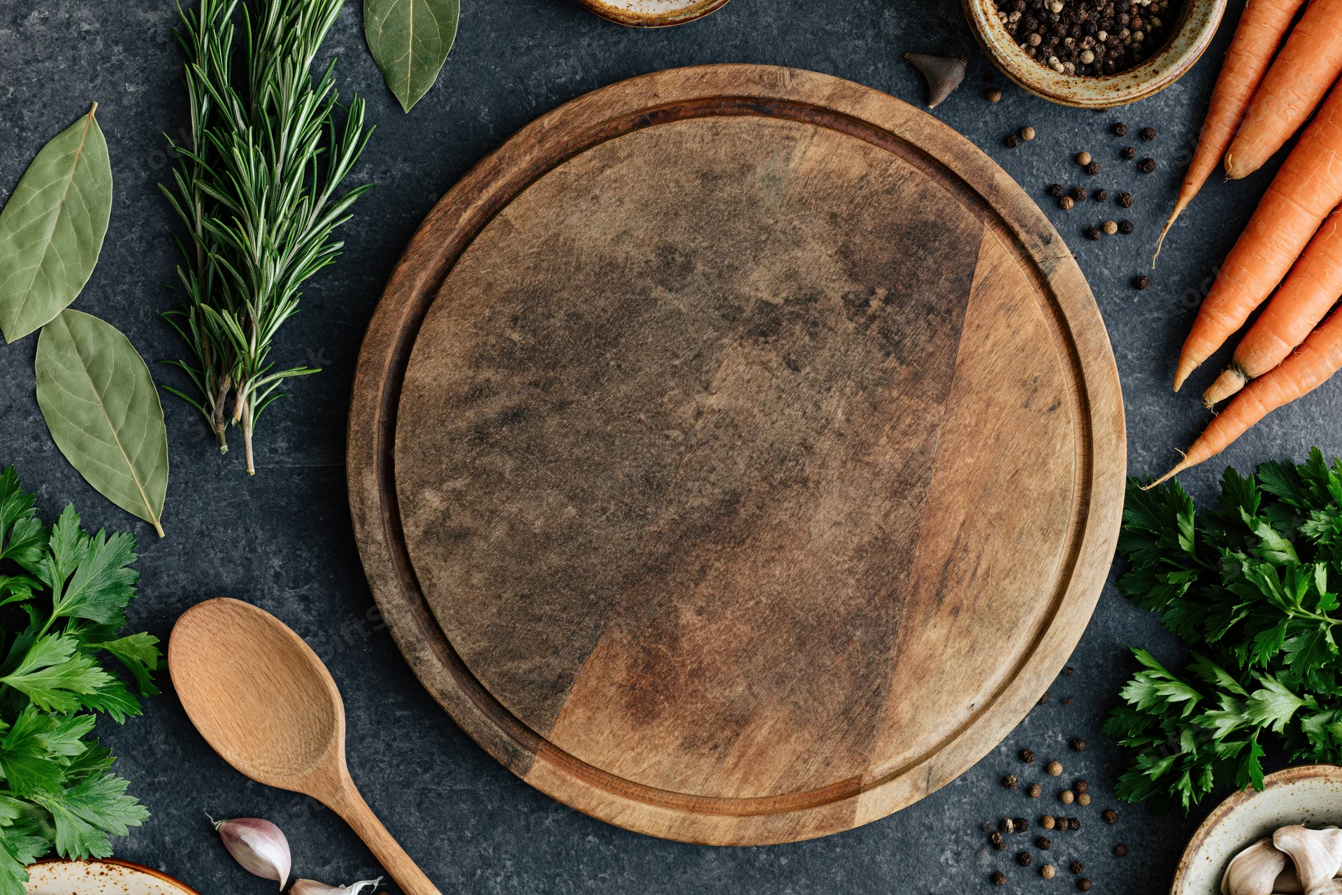 Round wooden cutting board surrounded by fresh herbs and vegetables on dark surface