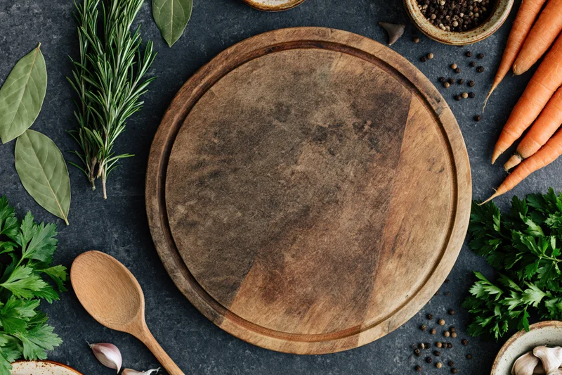 Round wooden cutting board surrounded by fresh herbs and vegetables on dark surface