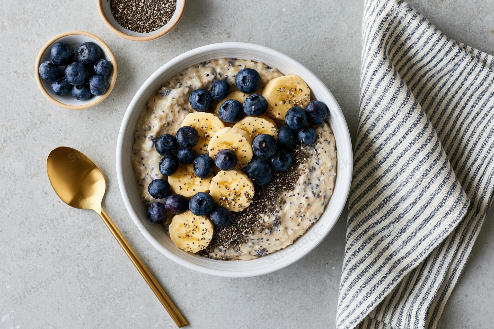 Creamy oatmeal topped with banana slices, blueberries and chia seeds in a white bowl on a gray countertop with a golden spoon and striped napkin beside it